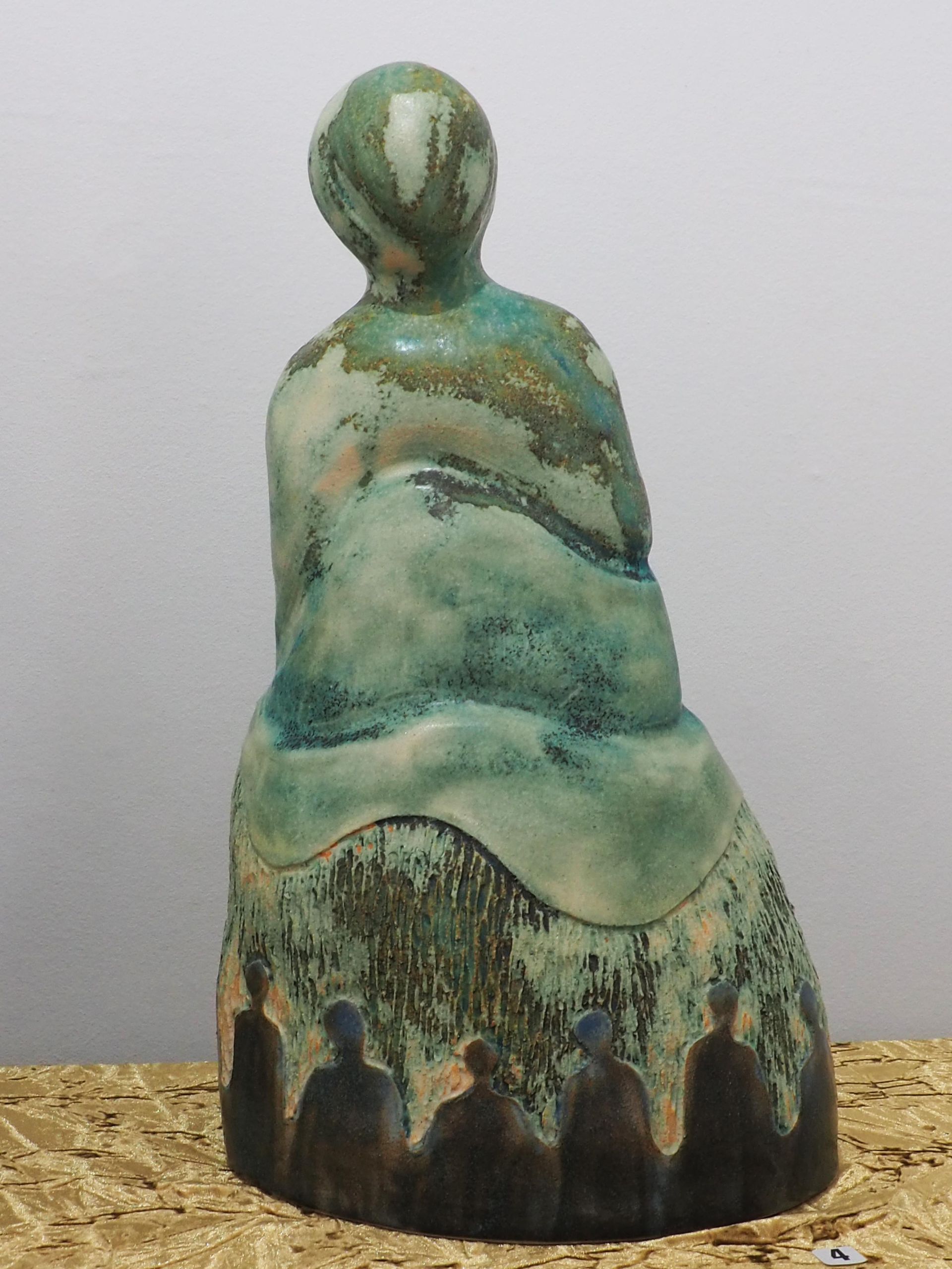 A sculpture of a smooth figure coming out of smooth ripling layers. The glaze is bronze brown at the bottom and greens towards the top. Skirting the bottom are figures of people against a rough texture