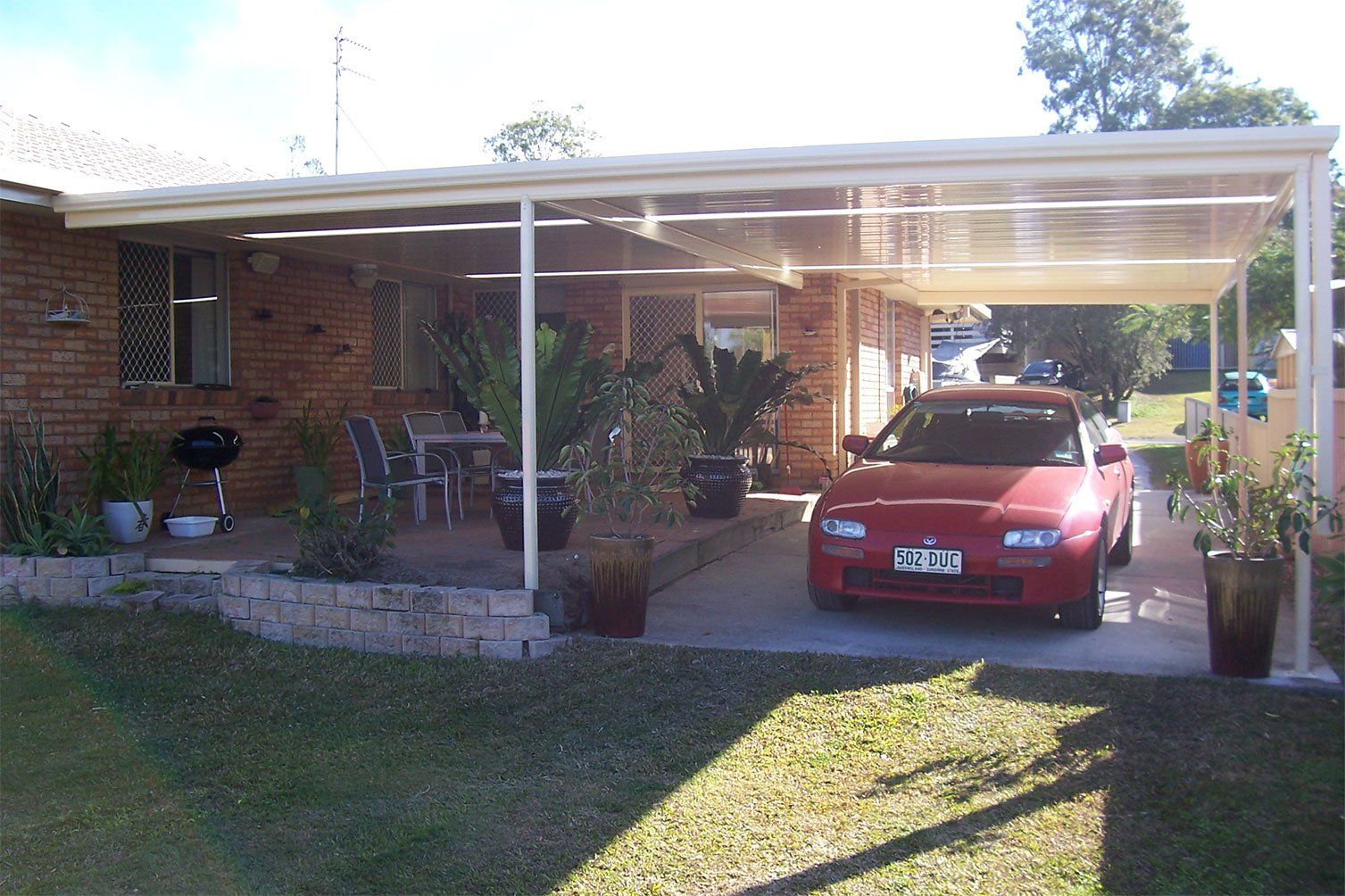 Red car parked under a carport beside a brick house, with potted plants and a grassy yard.