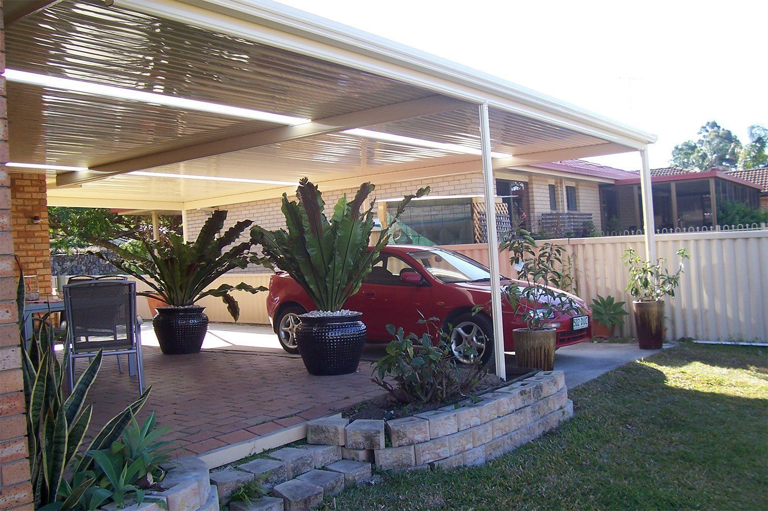 Red car parked under a carport beside potted plants and a white fence in a suburban yard.
