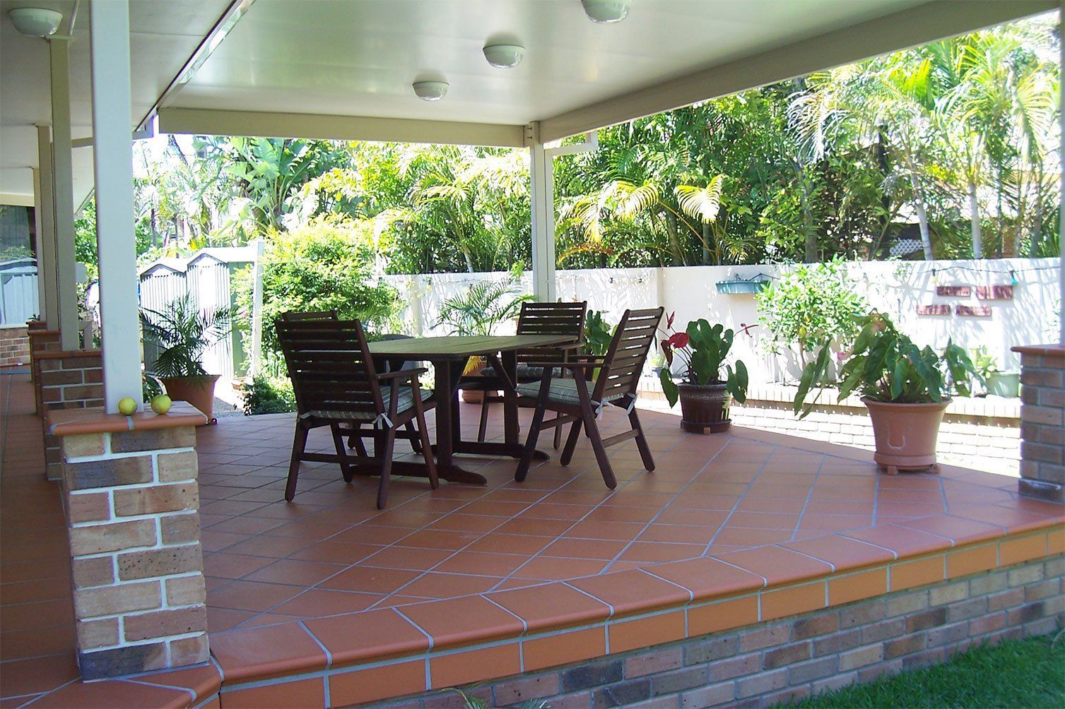 Covered patio with brick floor, wooden table and chairs, and potted plants overlooking a garden.