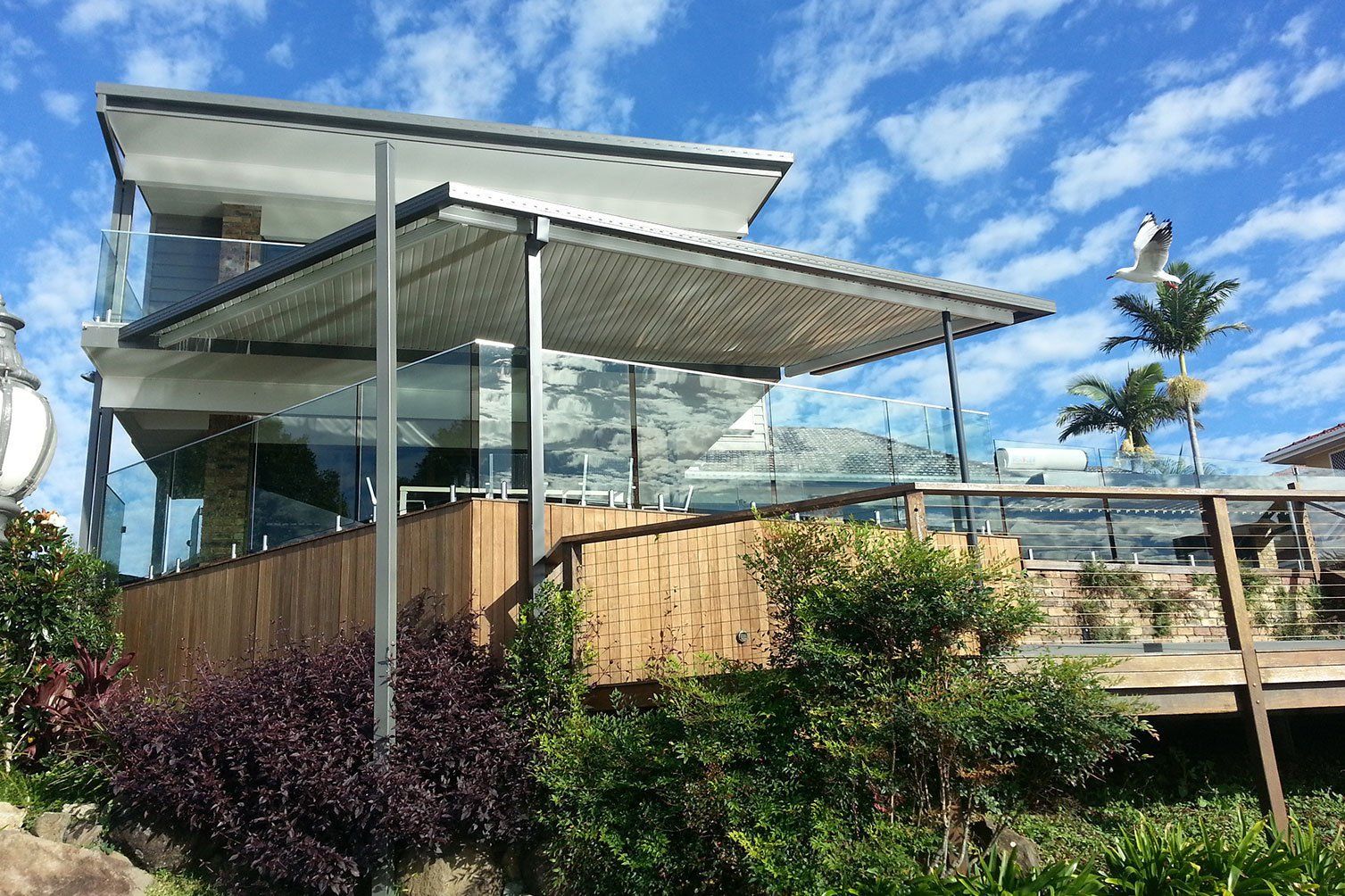 Modern glass-and-wood house with a wraparound deck under a blue sky and palm trees