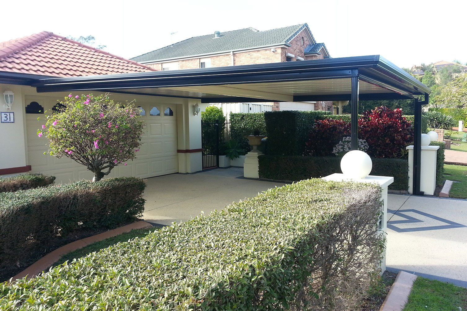 Covered driveway beside a stucco house, with manicured hedges and a tiled roof under bright sunlight