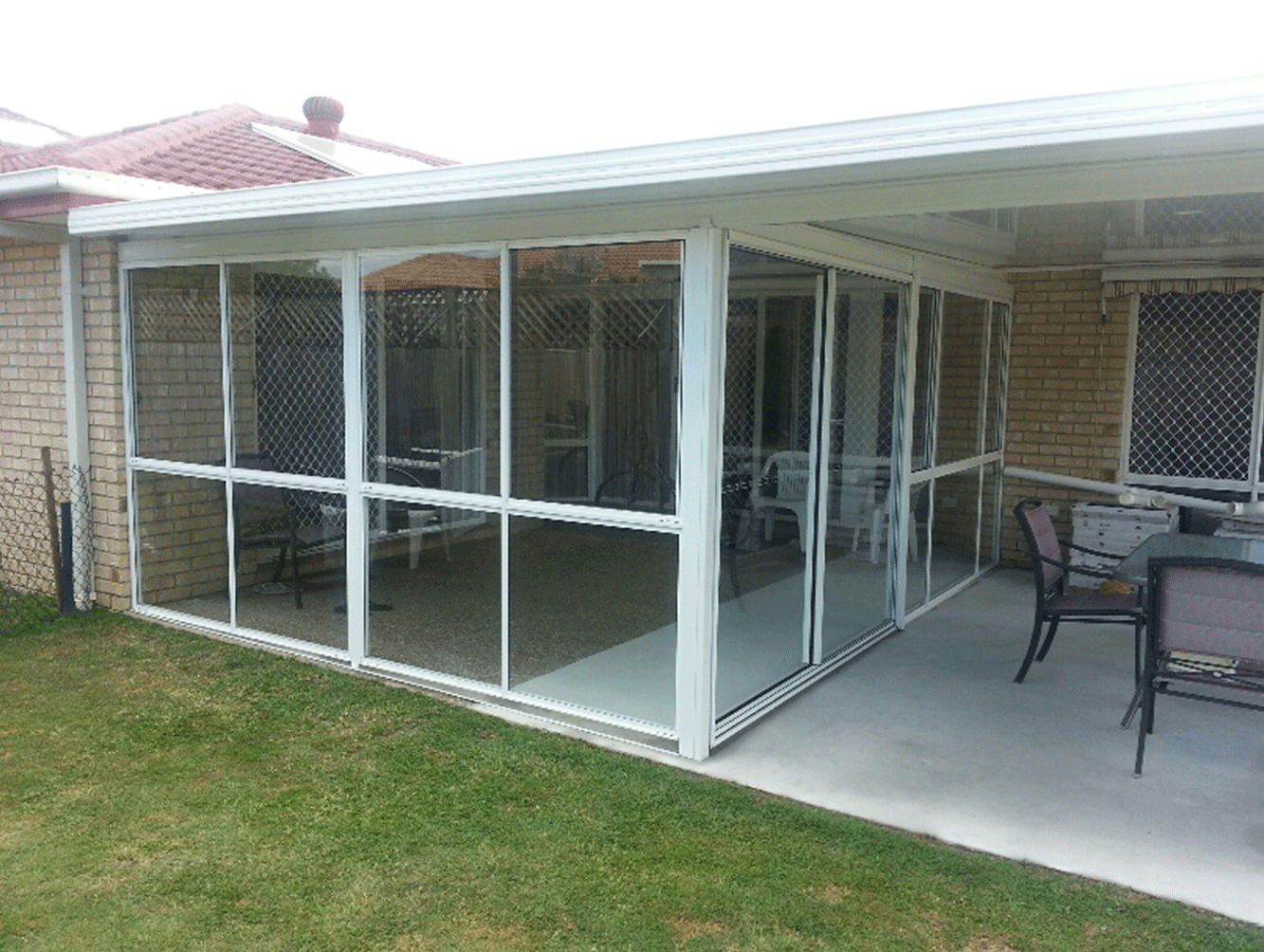 Screened patio enclosure beside a brick house, with glass walls and outdoor seating on a concrete slab.