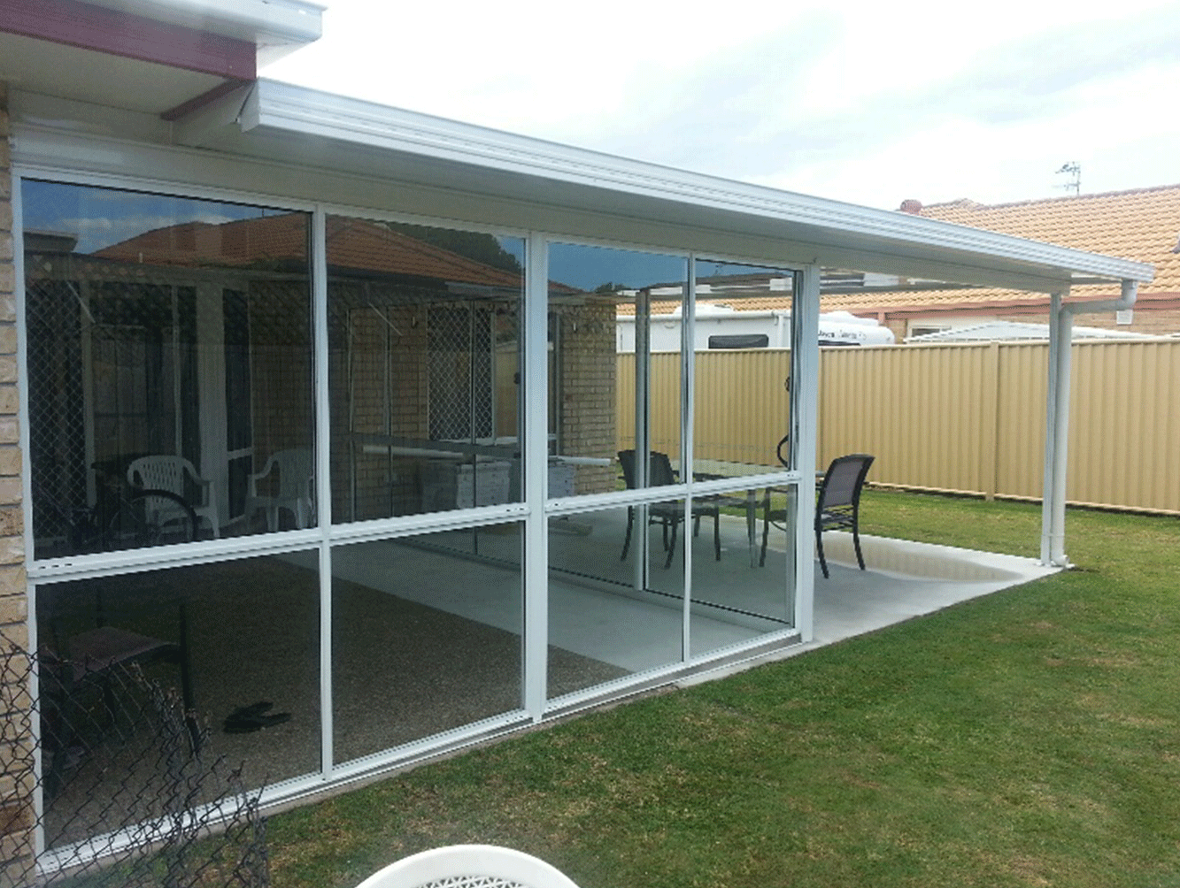 Covered patio with white-framed glass enclosure, patio chairs, and a lawn beside a fenced yard.