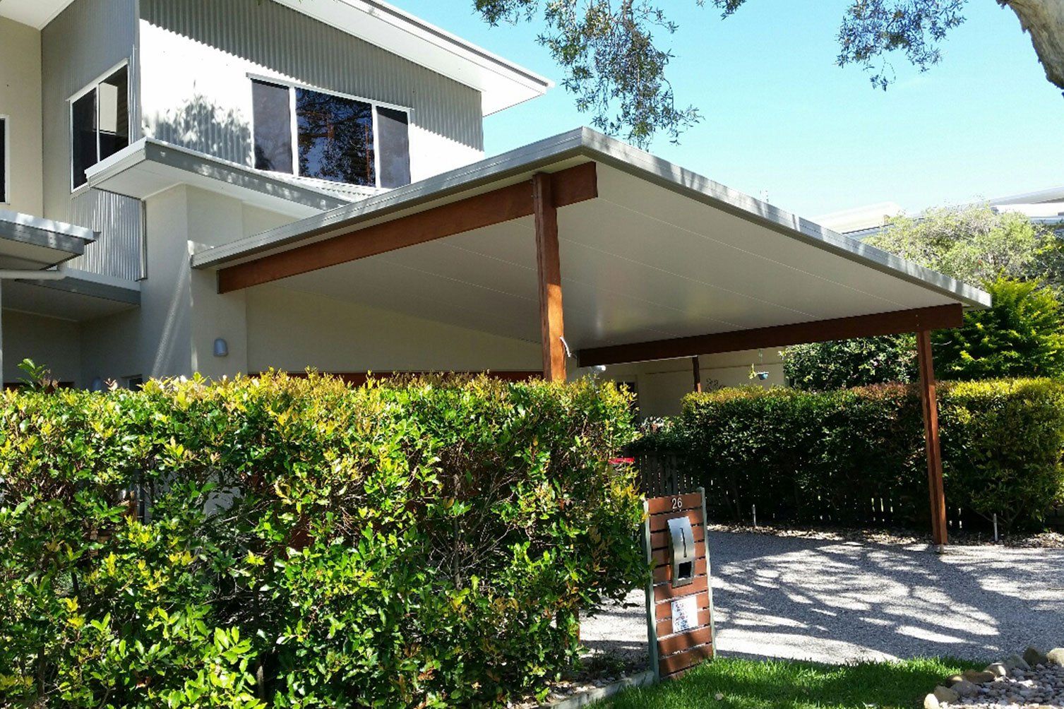 Modern house with a wooden carport roof, trimmed hedges, and a gravel driveway in sunlight