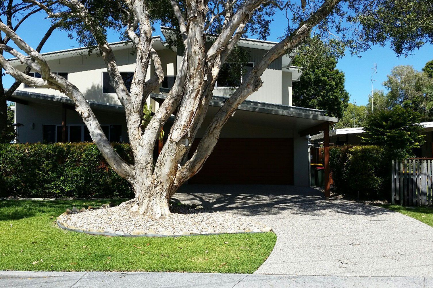 Two-story house with a large tree in the front yard and a gravel driveway under a clear blue sky