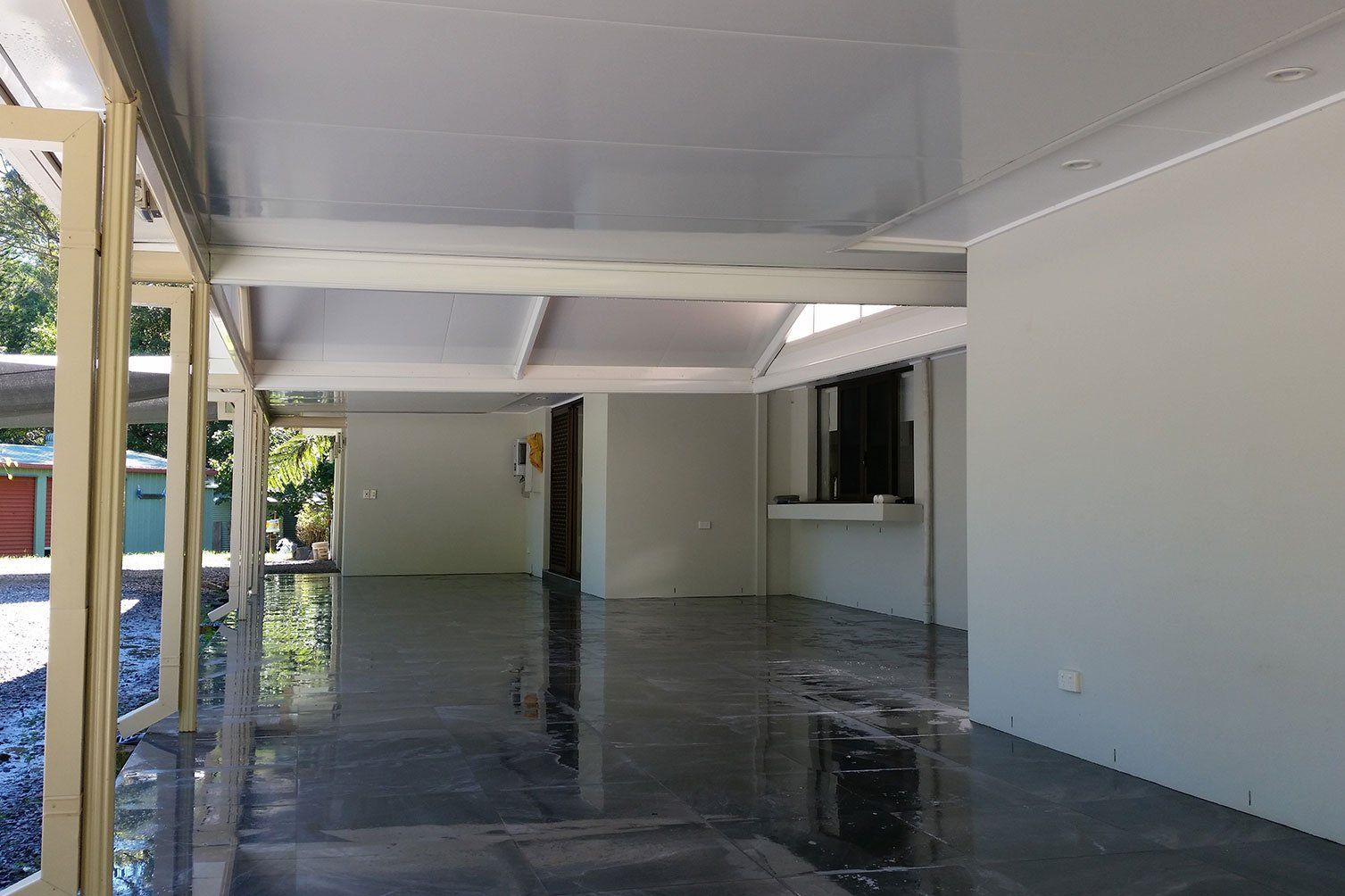 Covered patio with white walls, ceiling beams, and a glossy dark floor, opening to greenery outside