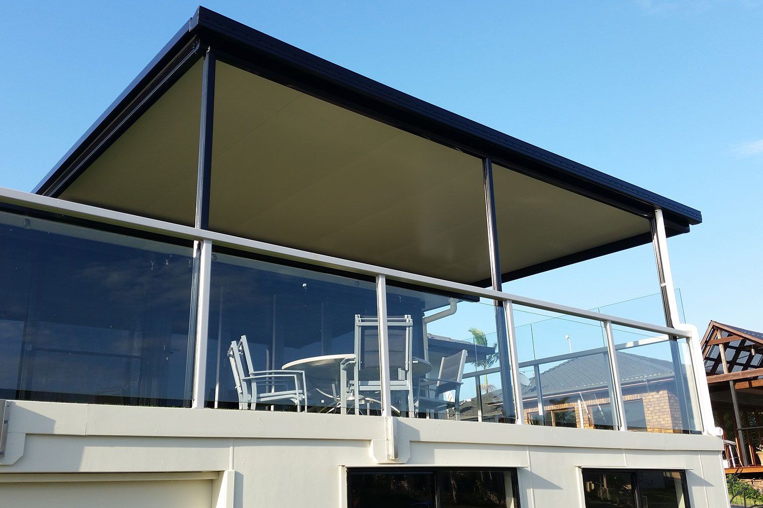Modern house balcony with glass railing and outdoor seating under a dark roof against a blue sky