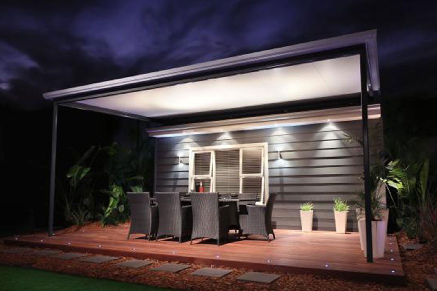 Covered patio with dining table and chairs at night, lit by warm lights beside a house