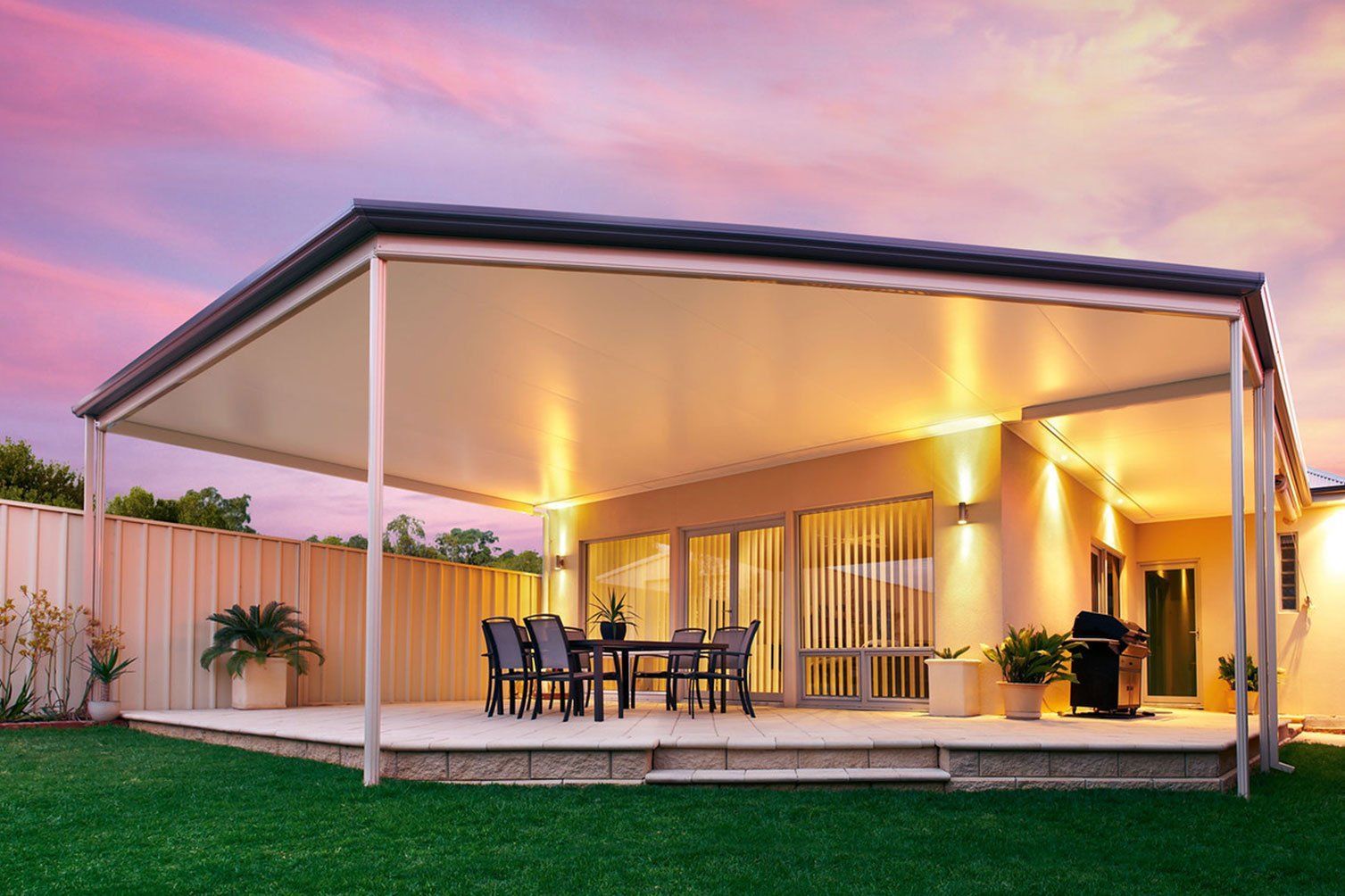 Covered patio with dining table and grill, lit warmly at sunset beside a fenced backyard.