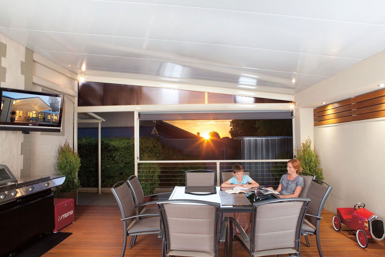 Covered patio with dining table, chairs, TV, and sunset view through open windows.