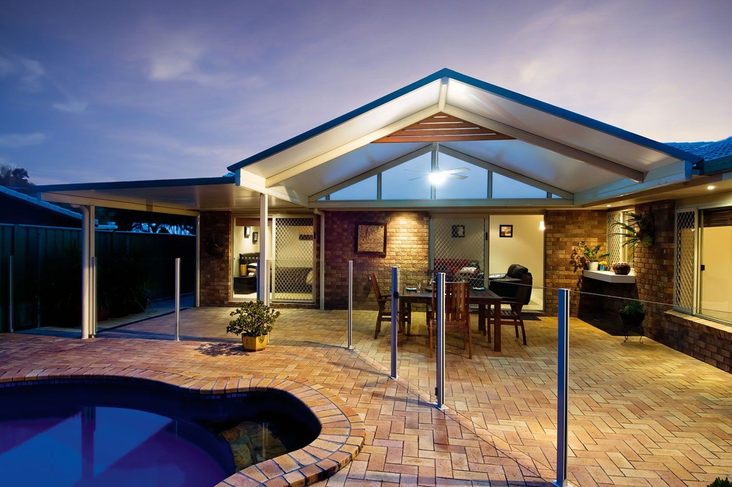 Covered backyard patio with pool at dusk, brick paving, outdoor dining table, and warm lights.