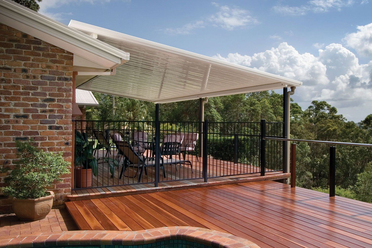 Covered wooden deck with outdoor seating beside a brick house, overlooking a green landscape under a cloudy sky
