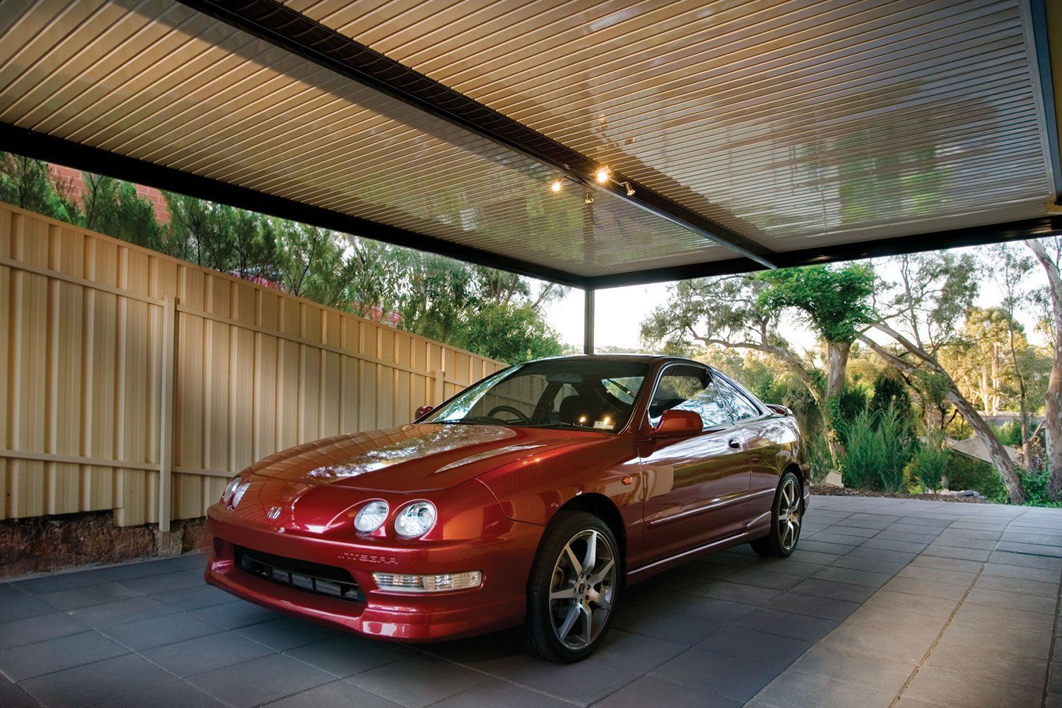Red sports car parked under a covered driveway beside a corrugated fence and trees