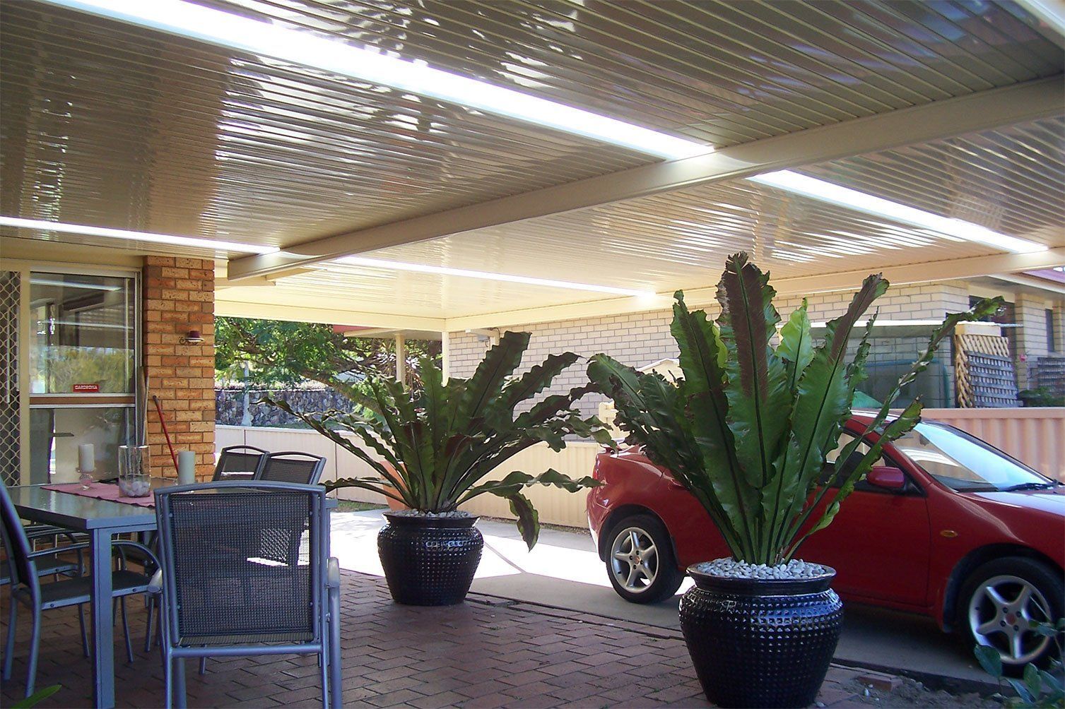 Patio with potted plants, outdoor chairs, and a red car under a covered roof