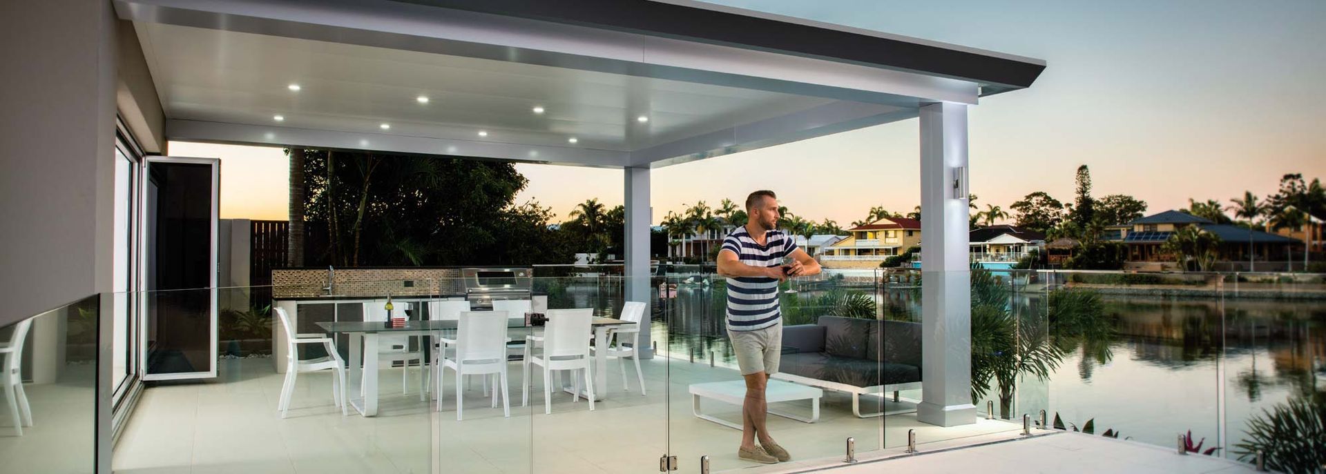 Modern patio with white furniture, glass railing, and a man standing by an open indoor-outdoor living area at sunset