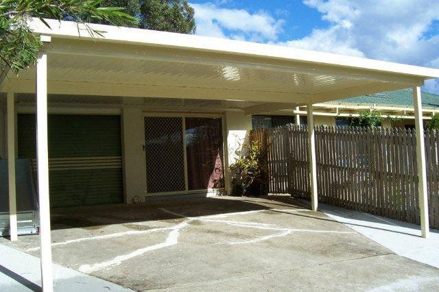 Covered driveway beside a house with a green garage door and wooden fence under a sunny sky