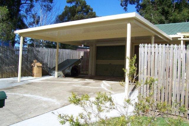Carport with a white roof beside a house, concrete driveway, and wooden fence in a sunny yard