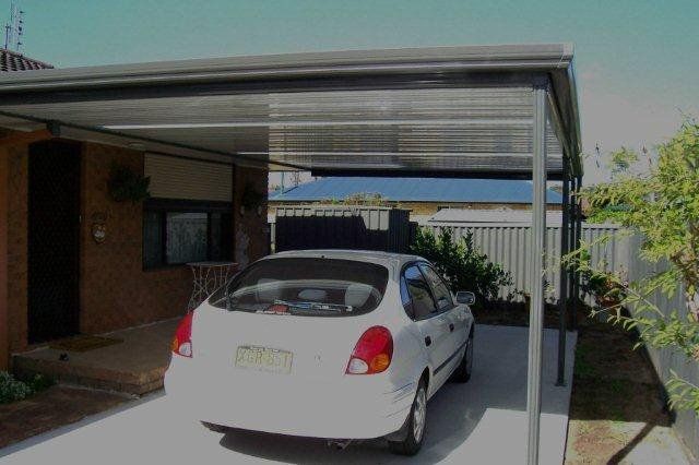 White car parked under a metal carport beside a brick house with a fence and garden.