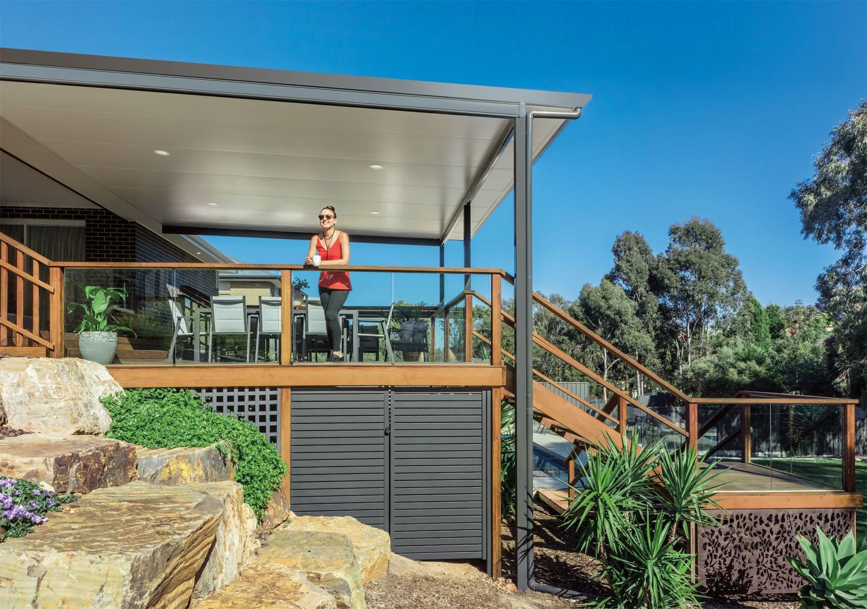 Person in red top standing on a deck with railing, under a roofed patio in a sunny backyard.