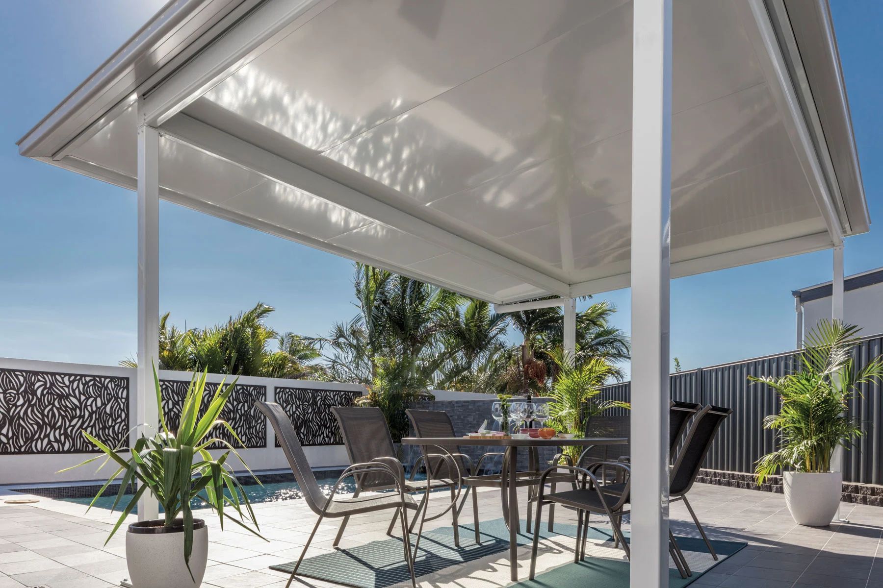 Covered patio with outdoor dining table and chairs under a white pergola in a sunny backyard.