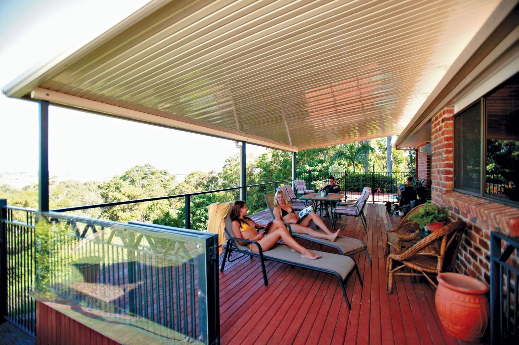 Covered outdoor deck with lounge chairs, red floorboards, and people relaxing beside a leafy view.