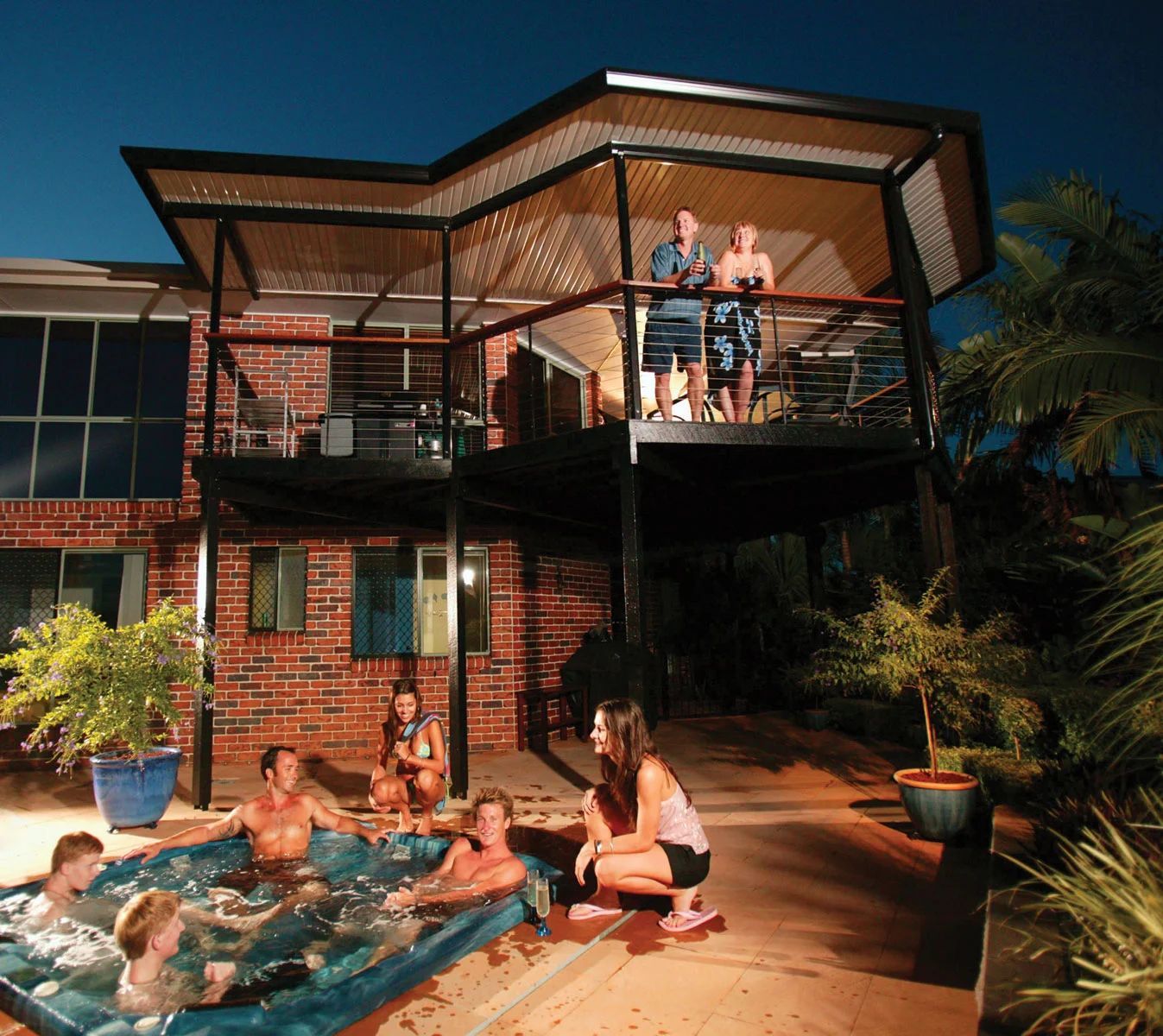 People relaxing around a backyard pool beside a brick house with an upper deck.