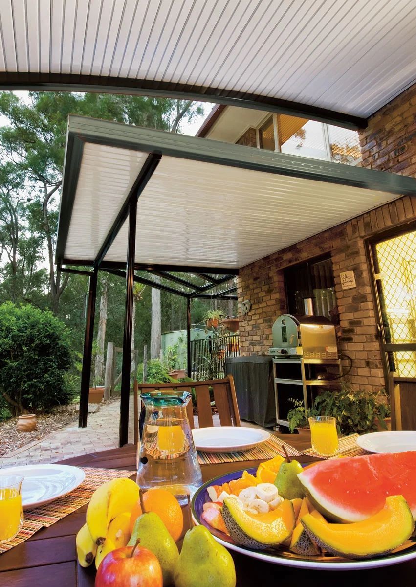 Outdoor dining table with fruit, juice, and plates beside a covered patio and garden seating area