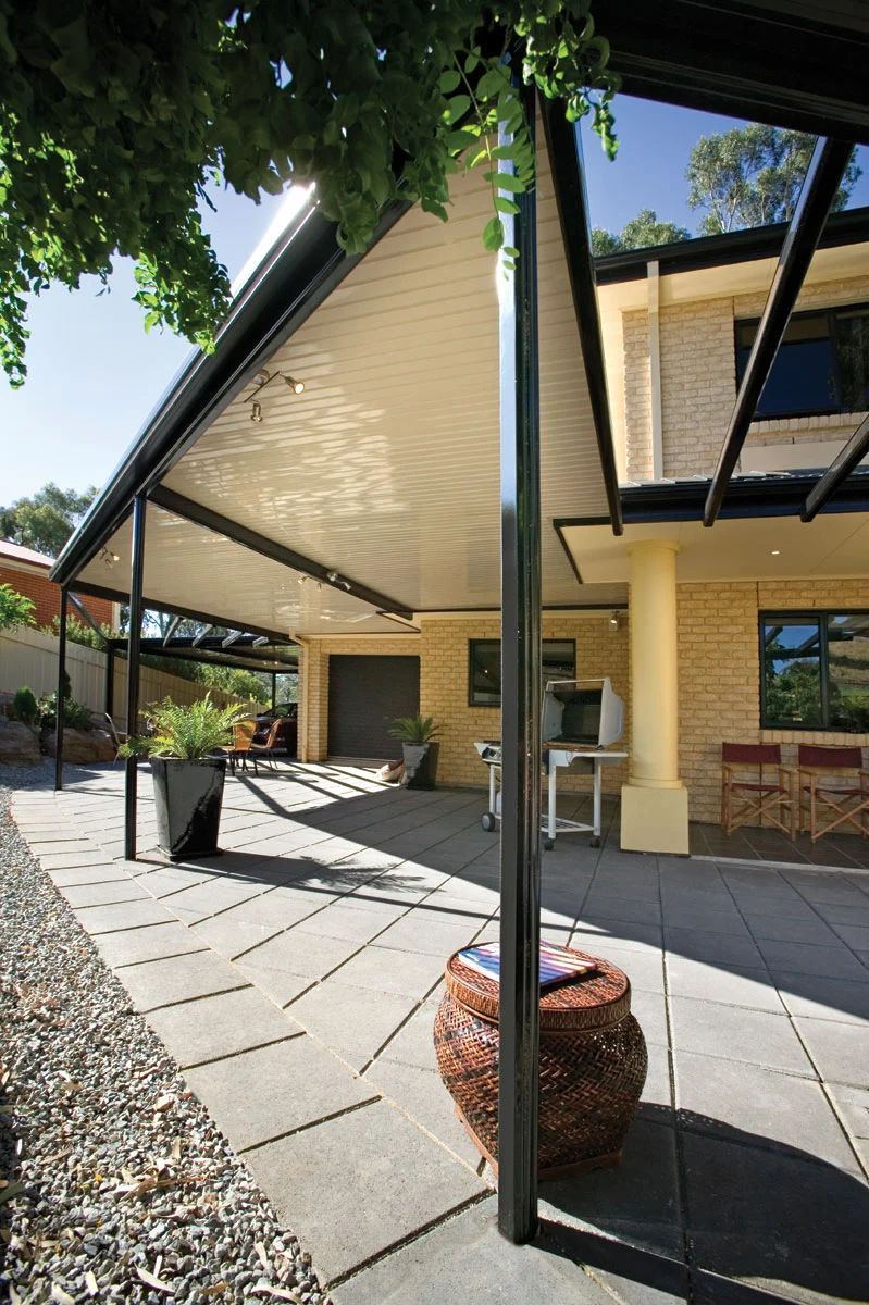 Covered patio with beige brick walls, black steel posts, and potted plants beside a house.