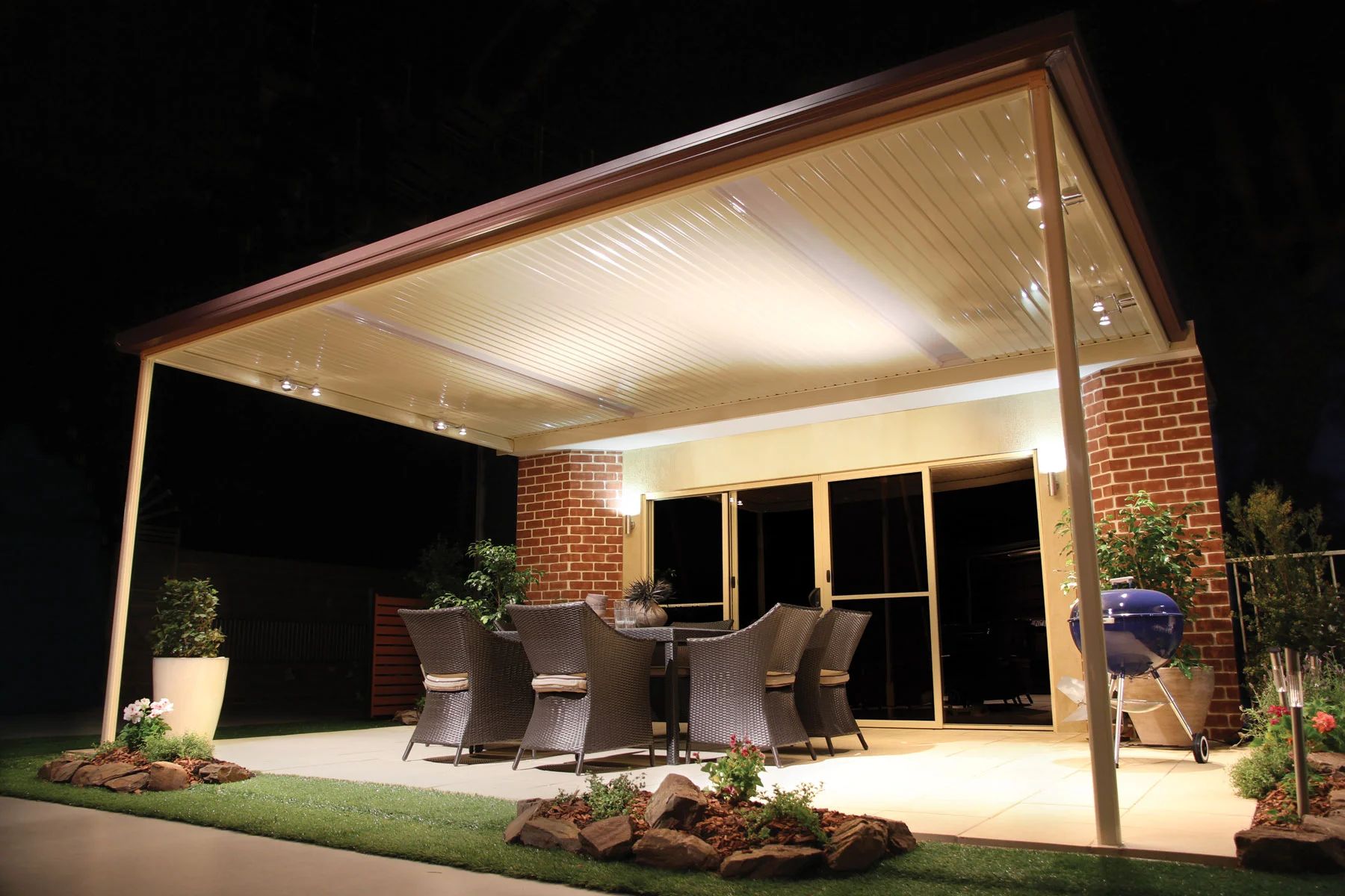 Covered patio with outdoor dining table and chairs, lit at night beside a brick house