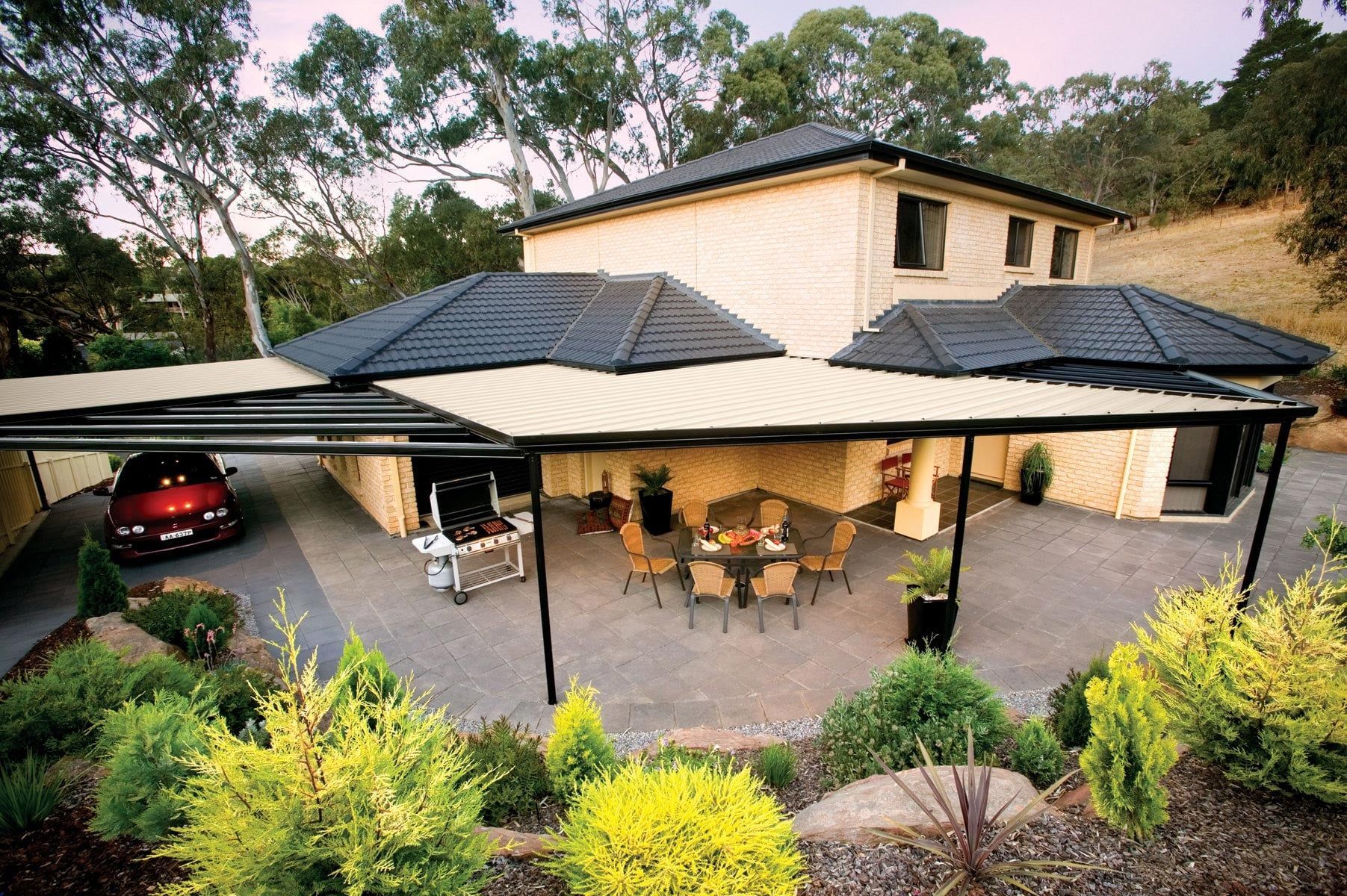 Modern house with a covered patio, outdoor seating, and a parked red car surrounded by greenery