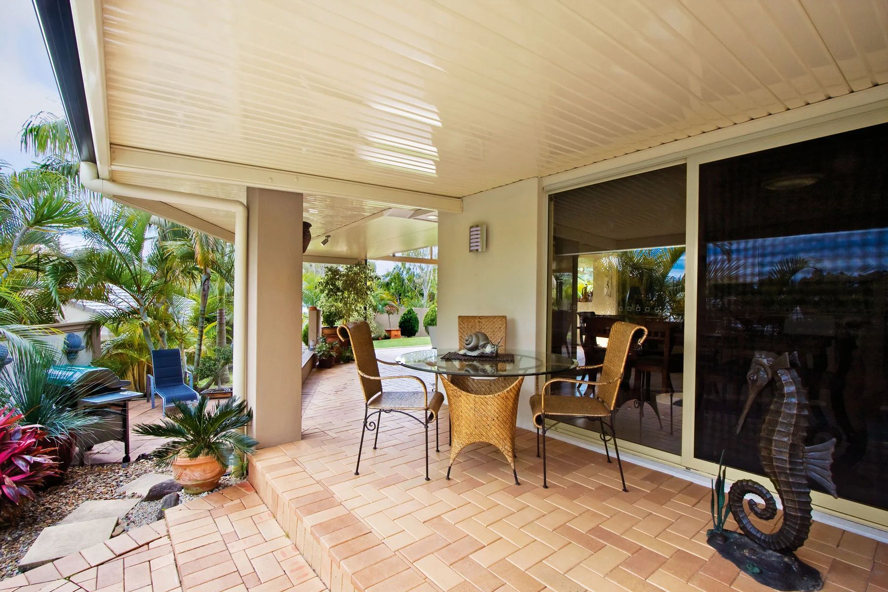 Covered patio with wicker chairs, glass table, and sliding doors overlooking a tropical garden.