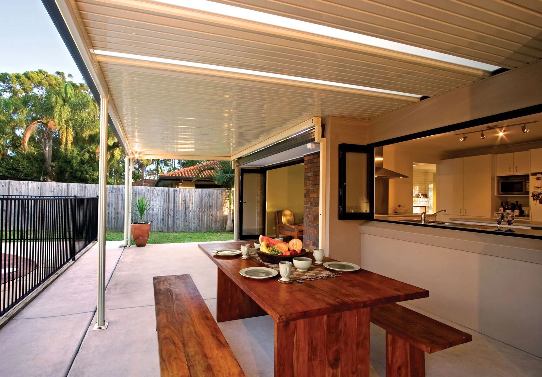 Covered patio with wooden picnic table beside a home kitchen, opening to a fenced backyard.