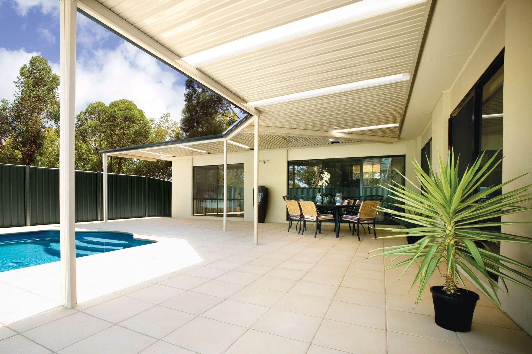 Covered patio beside a backyard pool, with white tiles, dining table, and potted plant.
