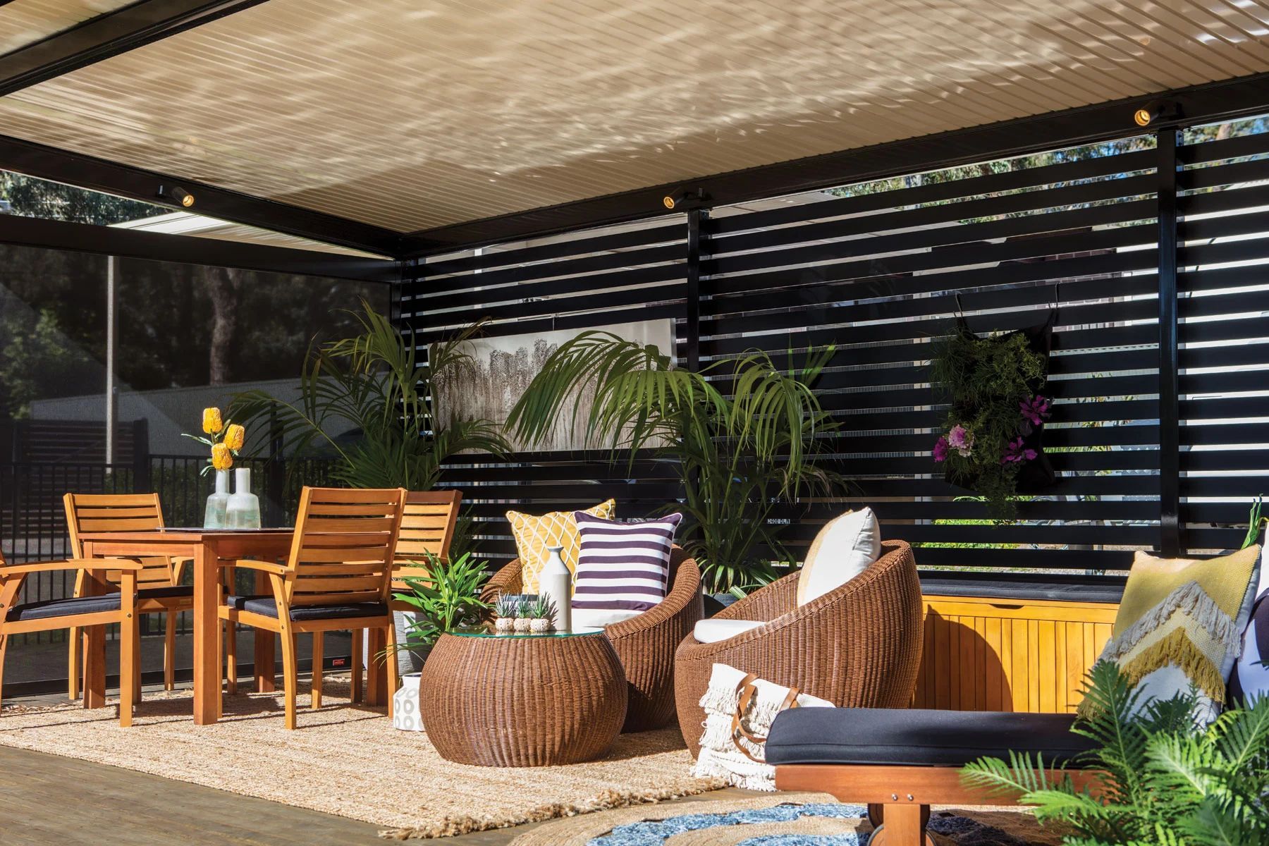 Stylish outdoor patio with wicker seating, wooden dining table, striped cushions, and lush plants under a slatted roof