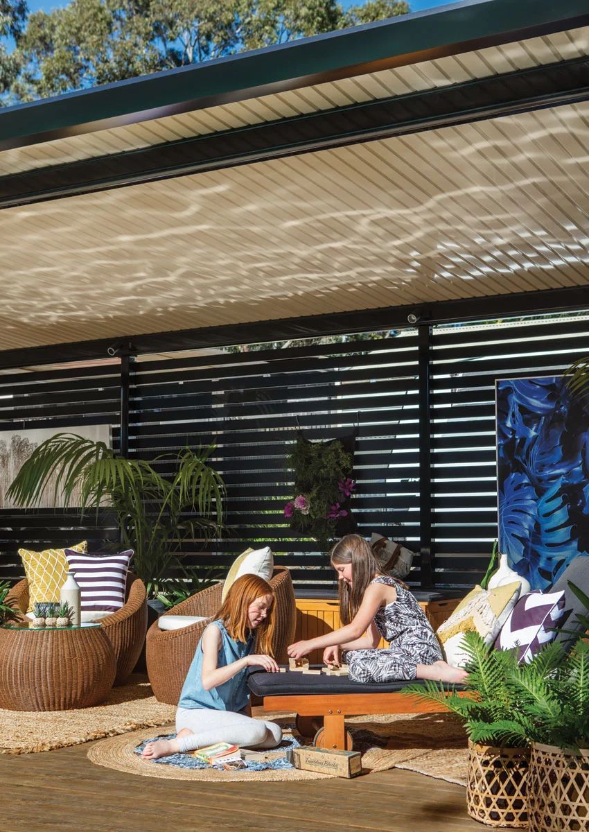 People relaxing on a sunlit patio with cushions, plants, and a low table under a shade awning