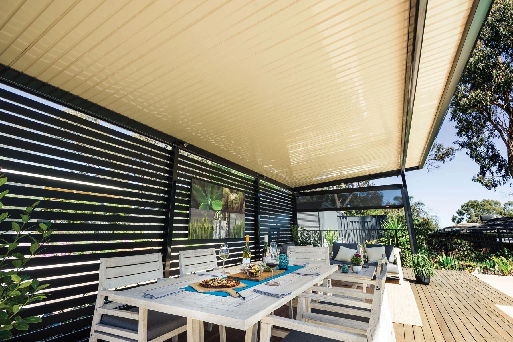 Covered outdoor dining area with white table and chairs on a wooden deck by greenery