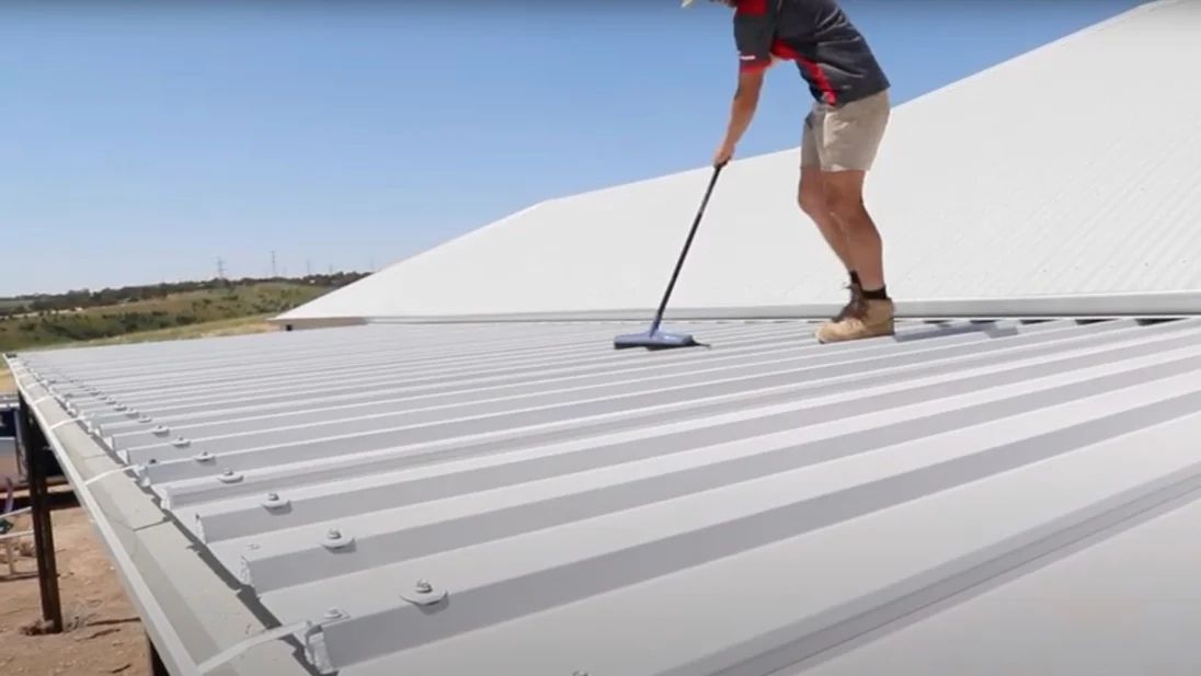 Person cleaning a white corrugated roof with a long-handled brush outdoors