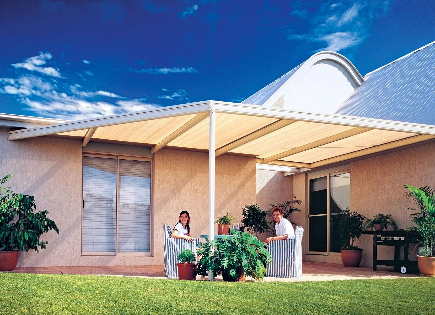 Two people standing on a covered patio beside a beige house with a blue roof and green lawn.
