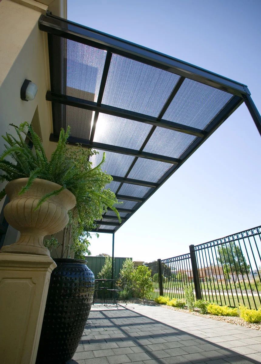 Covered patio walkway beside a house, with potted plants, metal railing, and bright sunlight.
