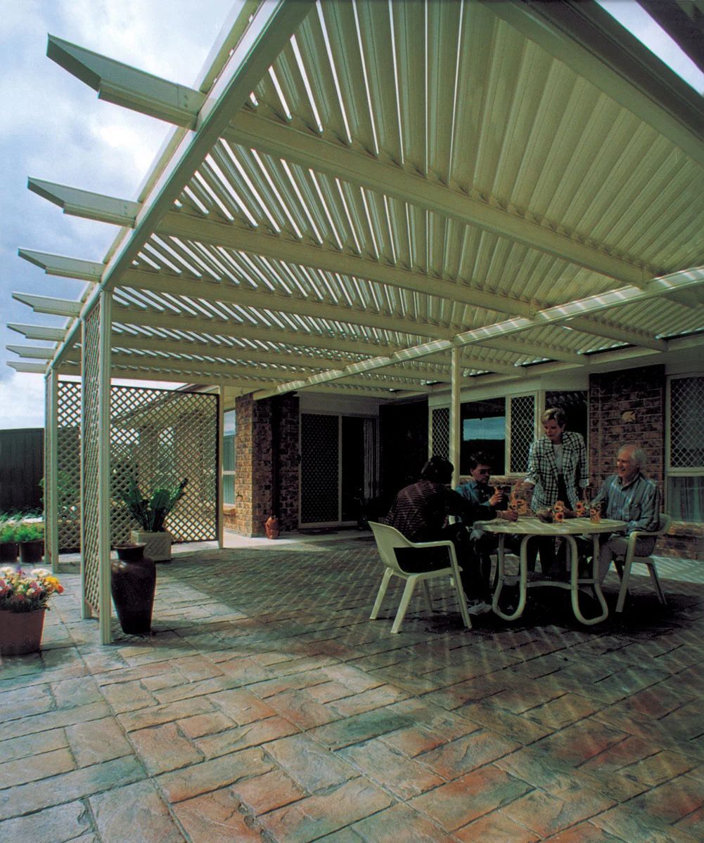Covered patio with pergola, stone flooring, and people seated at a table outside a brick house.