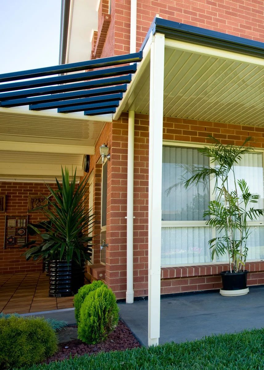 Covered porch with white posts, blue awning, brick wall, and potted plants by a window.