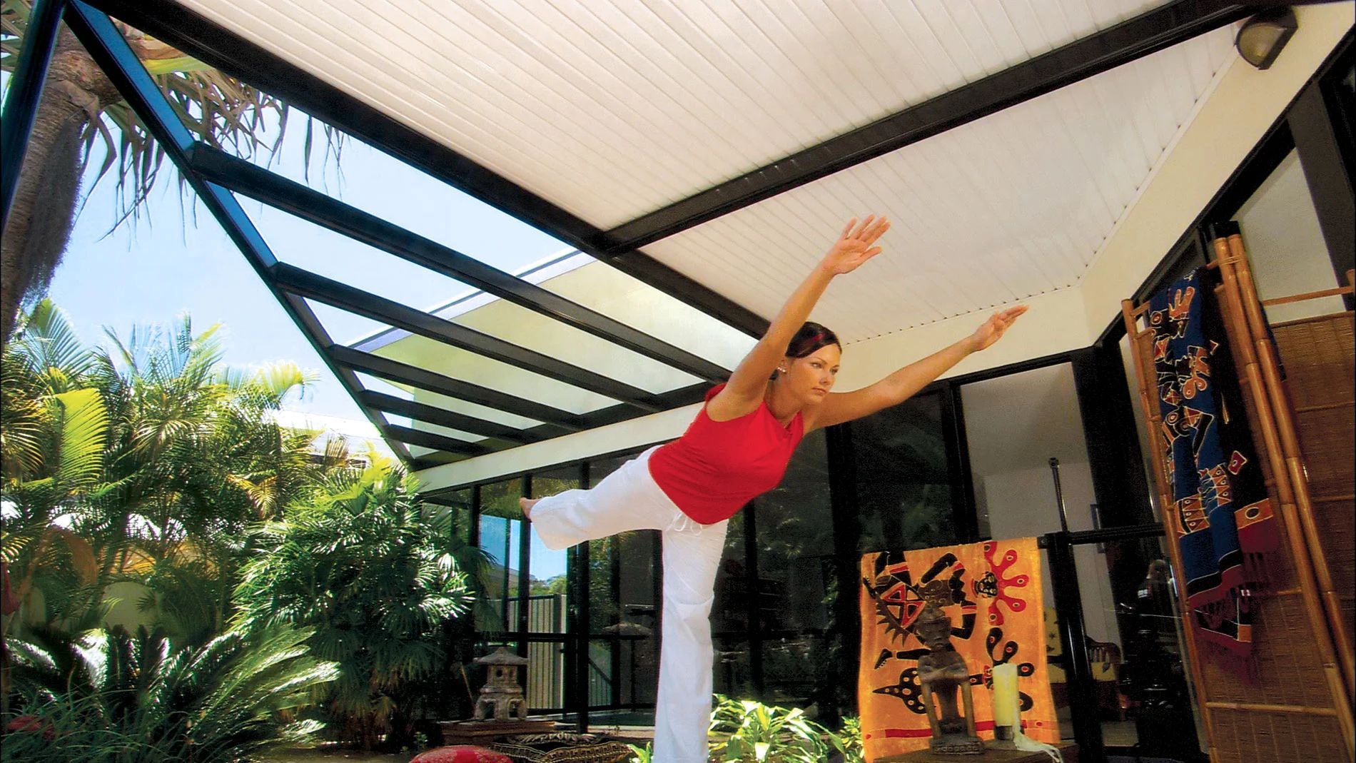 Person in pink top and white pants doing a yoga pose under a covered patio with tropical plants.