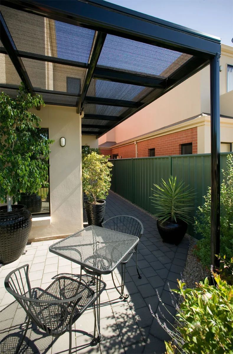 Patio with metal table and chairs under a black pergola beside potted plants and a green fence