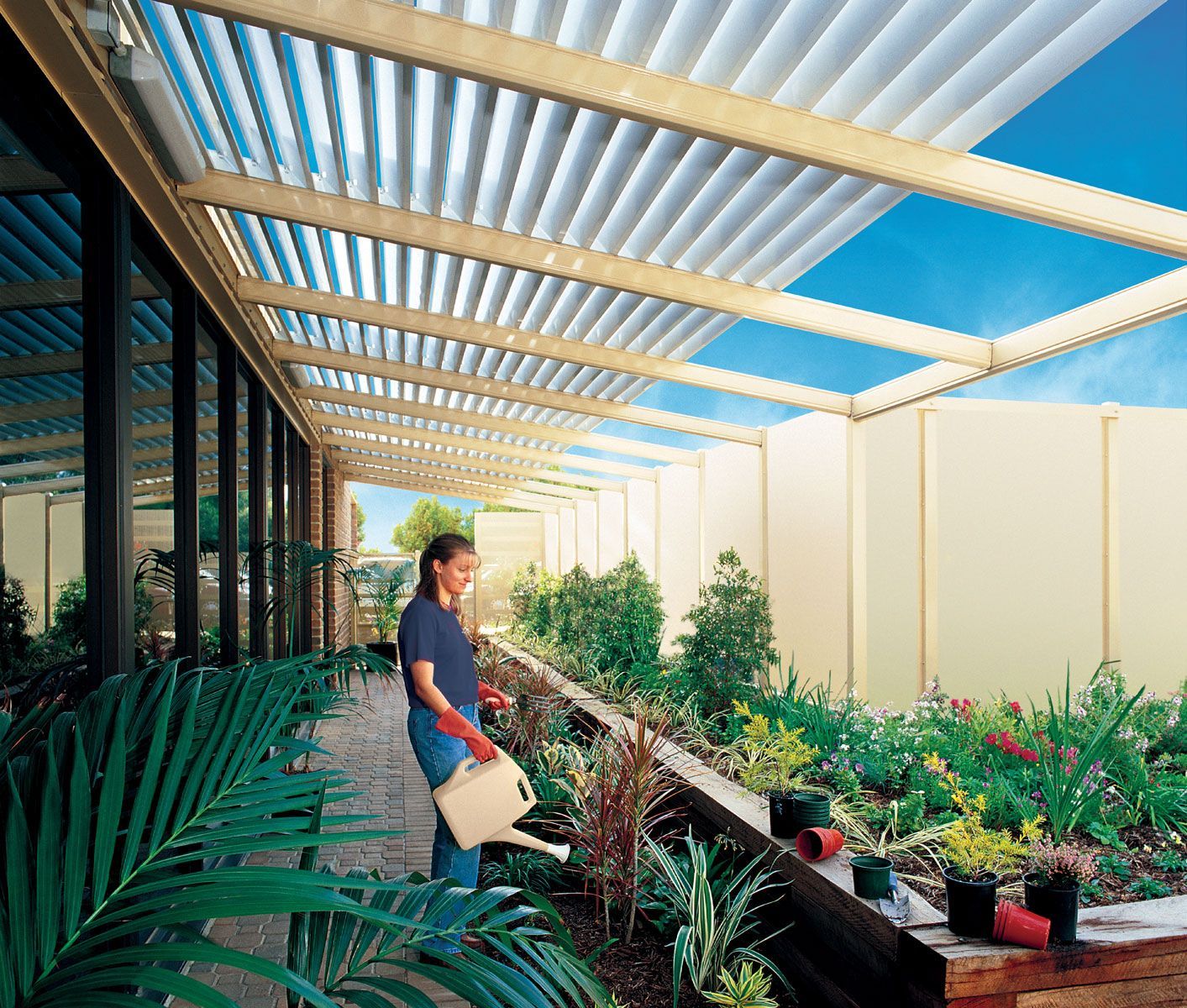 Person standing in a sunny garden walkway beside raised planters and lush green plants under a striped pergola