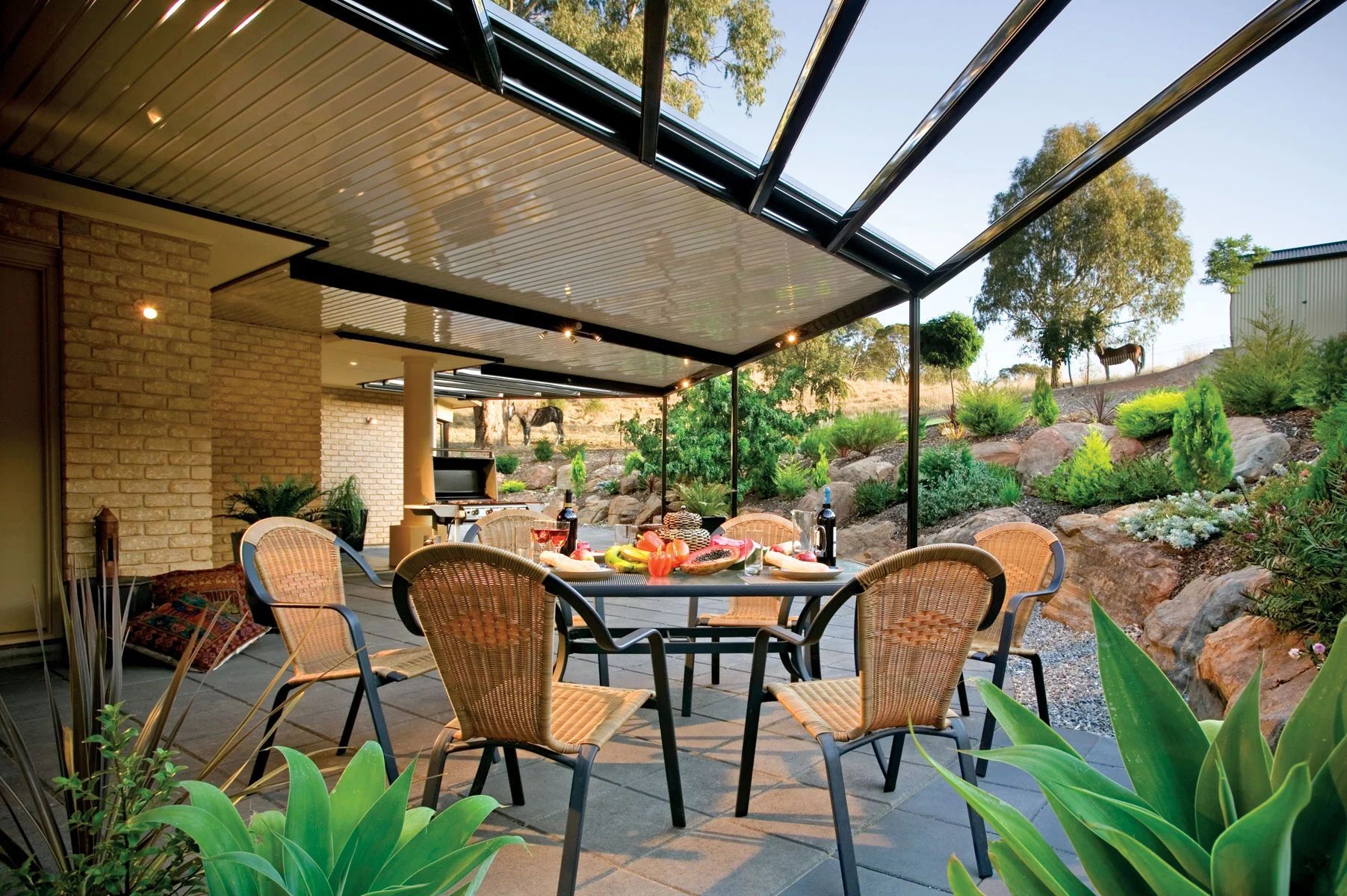 Outdoor patio dining area with wicker chairs, table setting, and pergola overlooking a landscaped garden