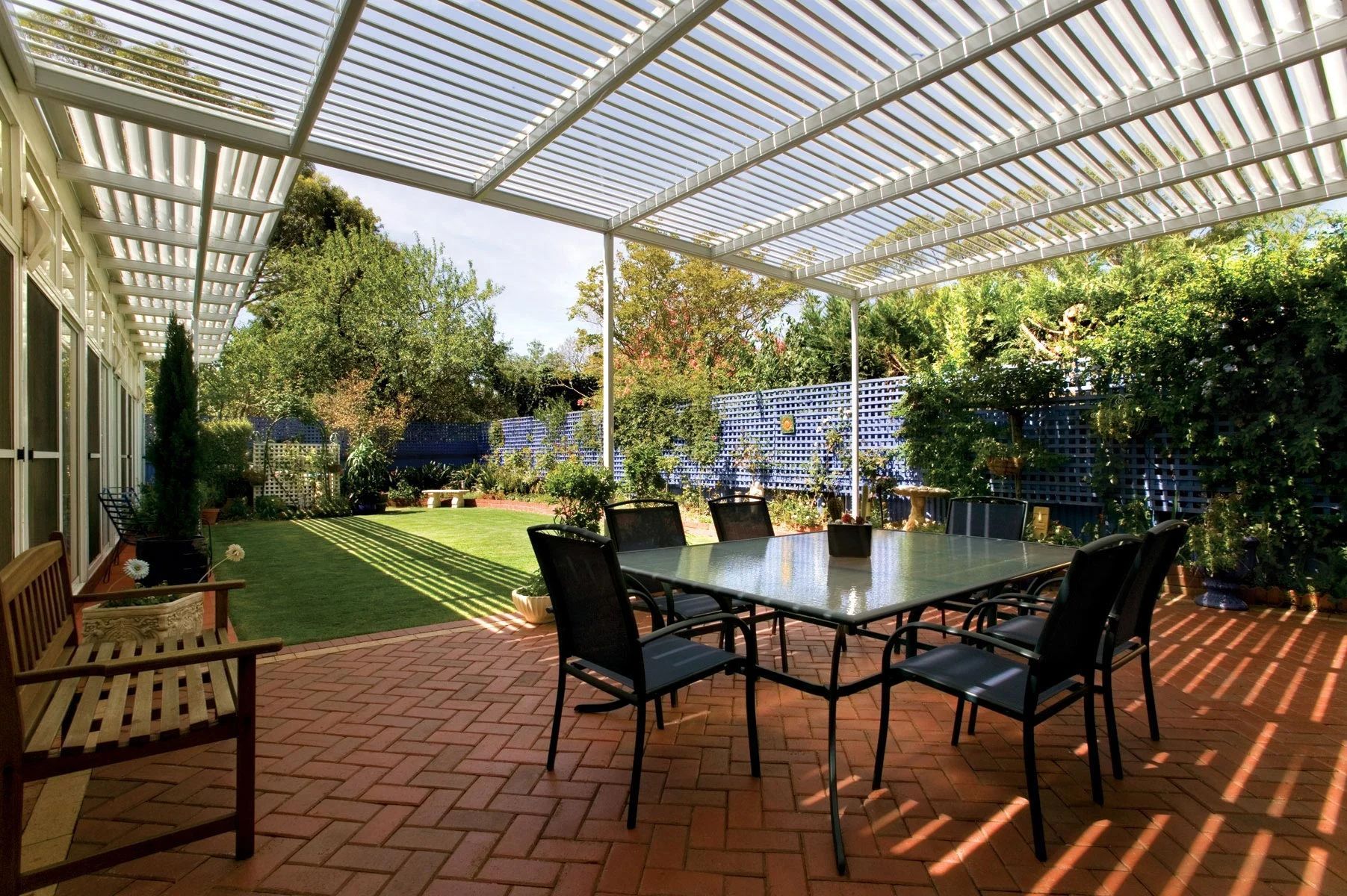 Patio with glass table and black chairs under a white pergola, surrounded by greenery and sunlight