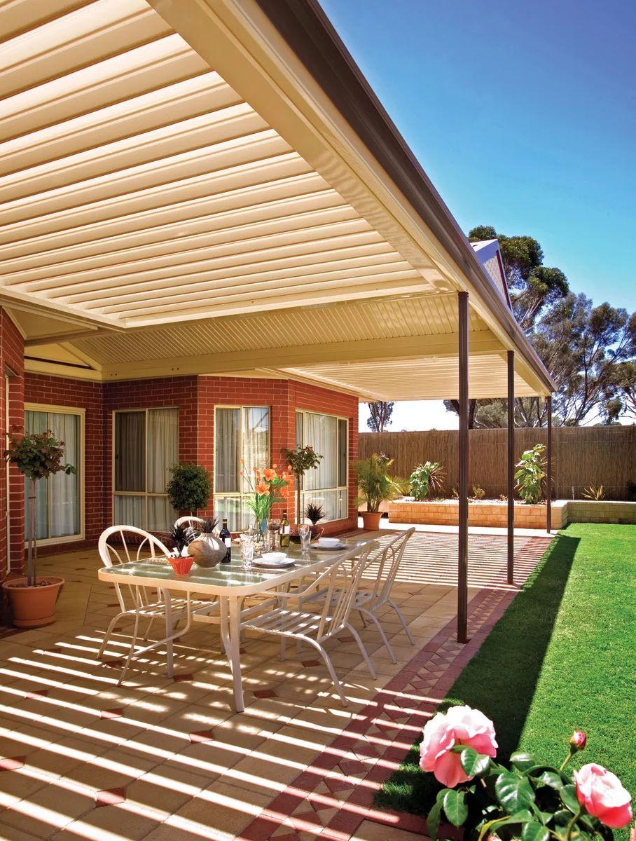 Covered patio with outdoor dining set, striped shade, potted flowers, and lawn beside a brick house
