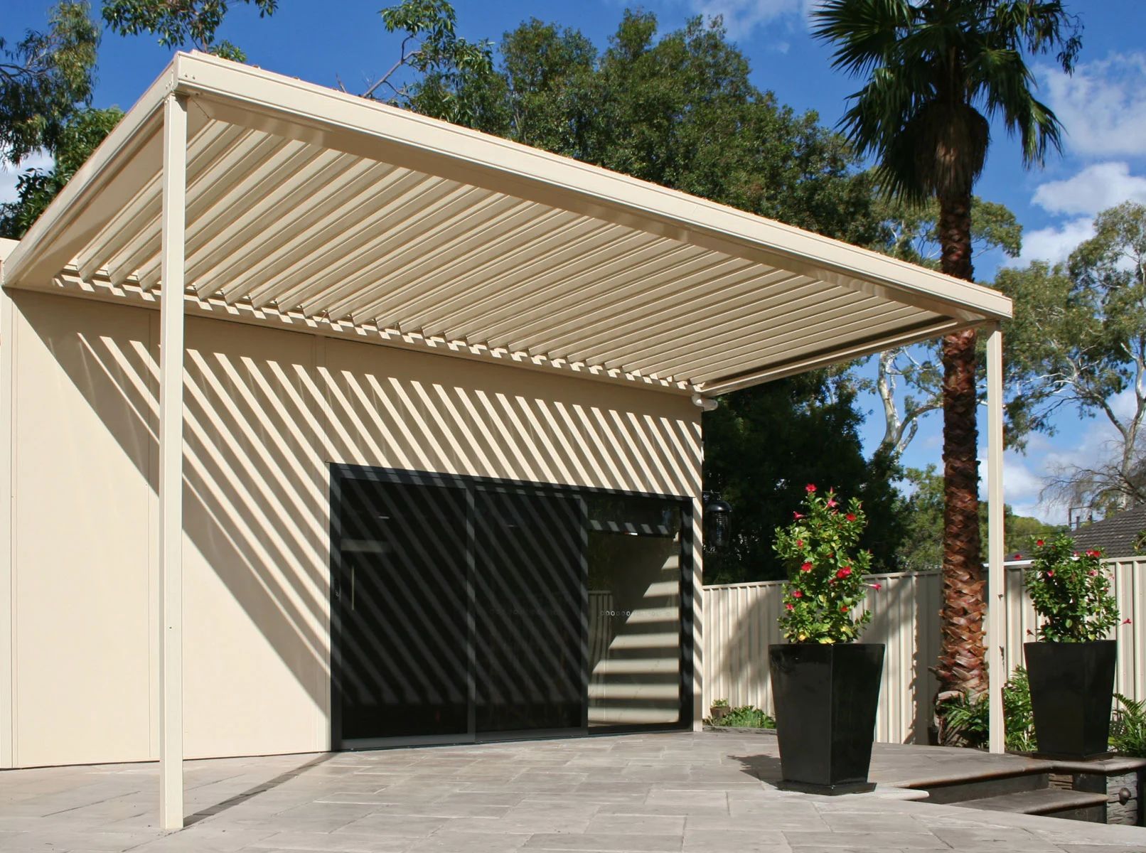 Modern carport with beige slatted roof, garage doors, and potted plant in a sunny courtyard