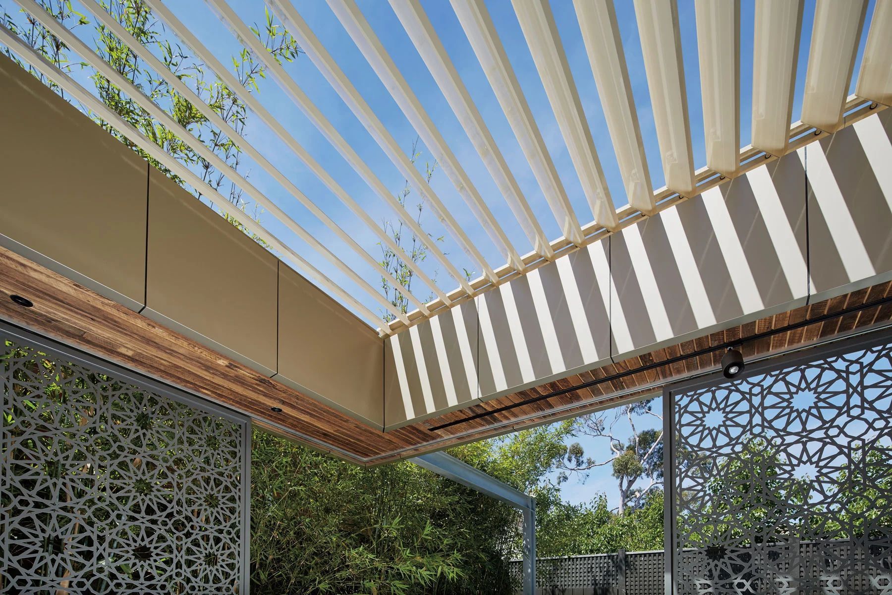 Outdoor patio with white pergola slats casting striped shadows over patterned walls and greenery.